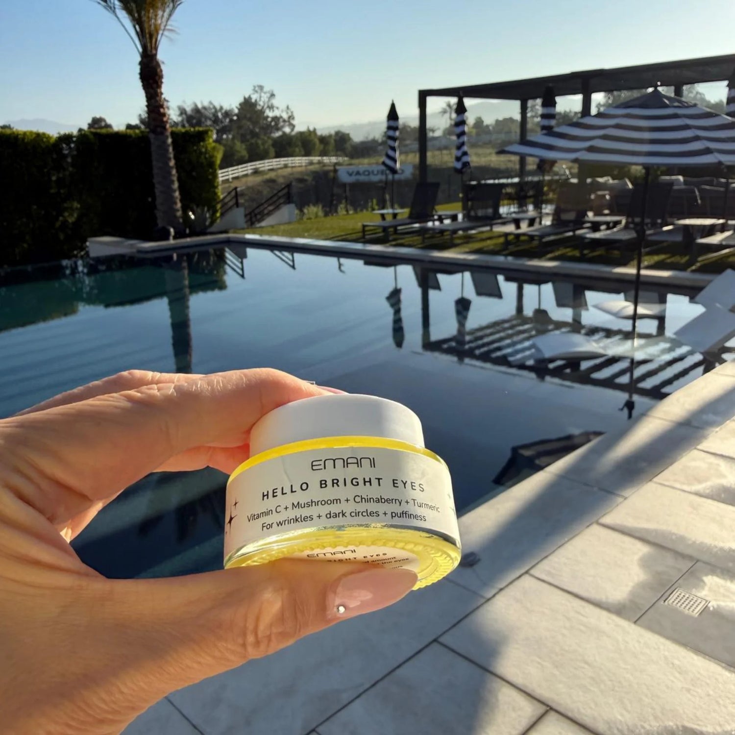 Hand holding a jar of Emani Hello Bright Eyes product by a poolside.