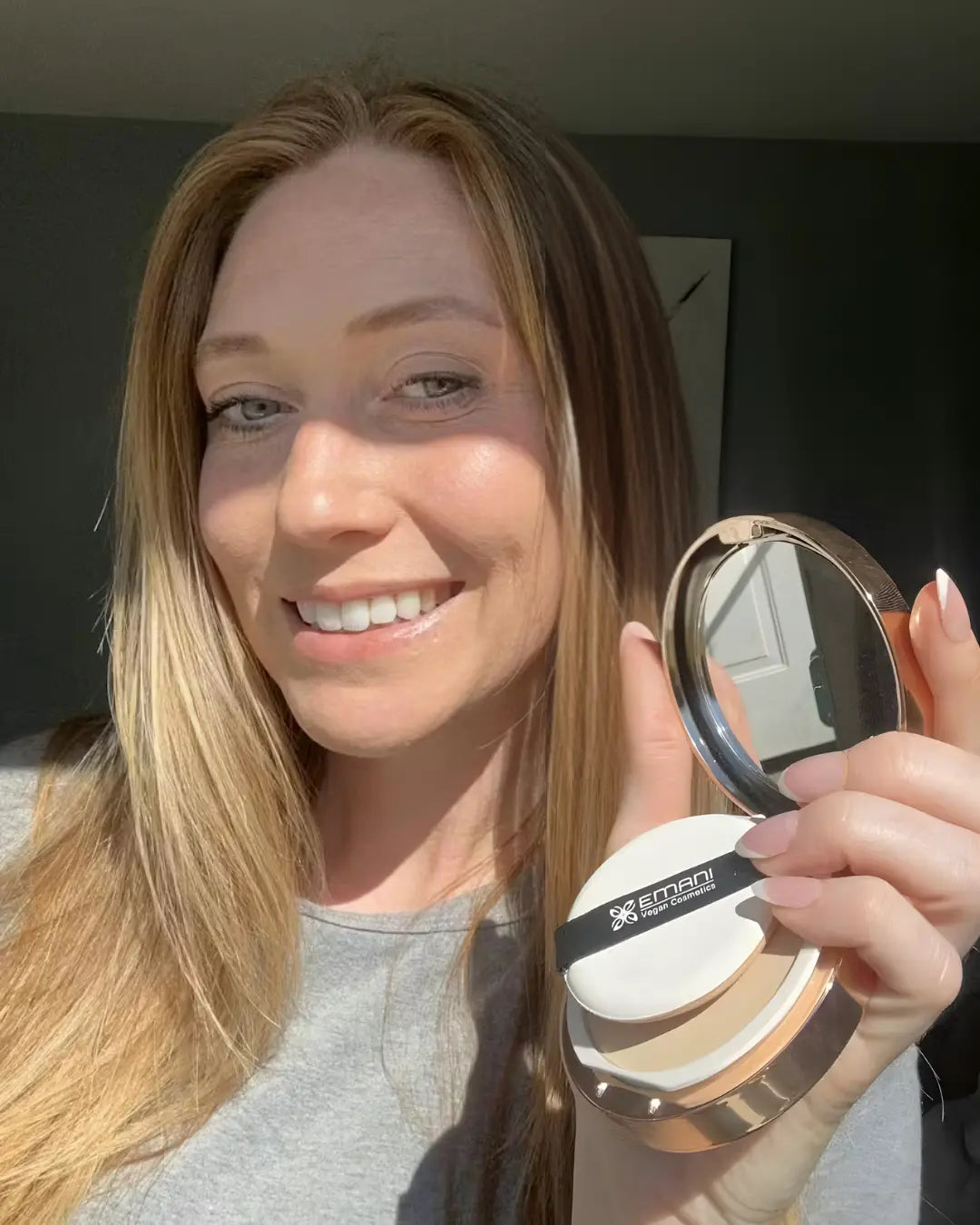 Woman holding a compact mirror with a brand logo, smiling outdoors. | Kody Hattendorf